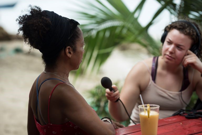 Person seated at picnic table wearing headphones holds a microphone in front of another person seated with their back to the camera. A palm tree and beach are in the background.