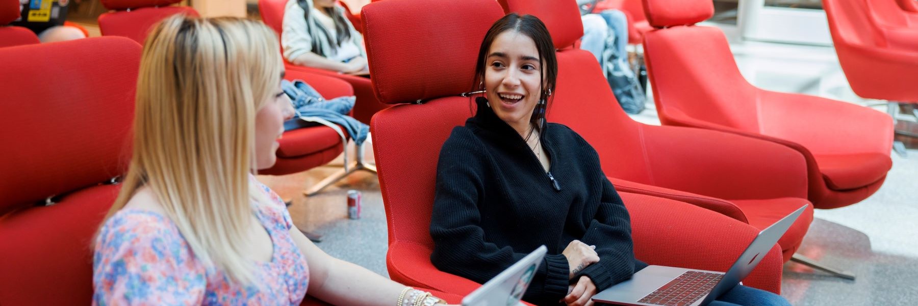 Two people are speaking to one another while seated in large, plush red chairs with laptops open on their laps.