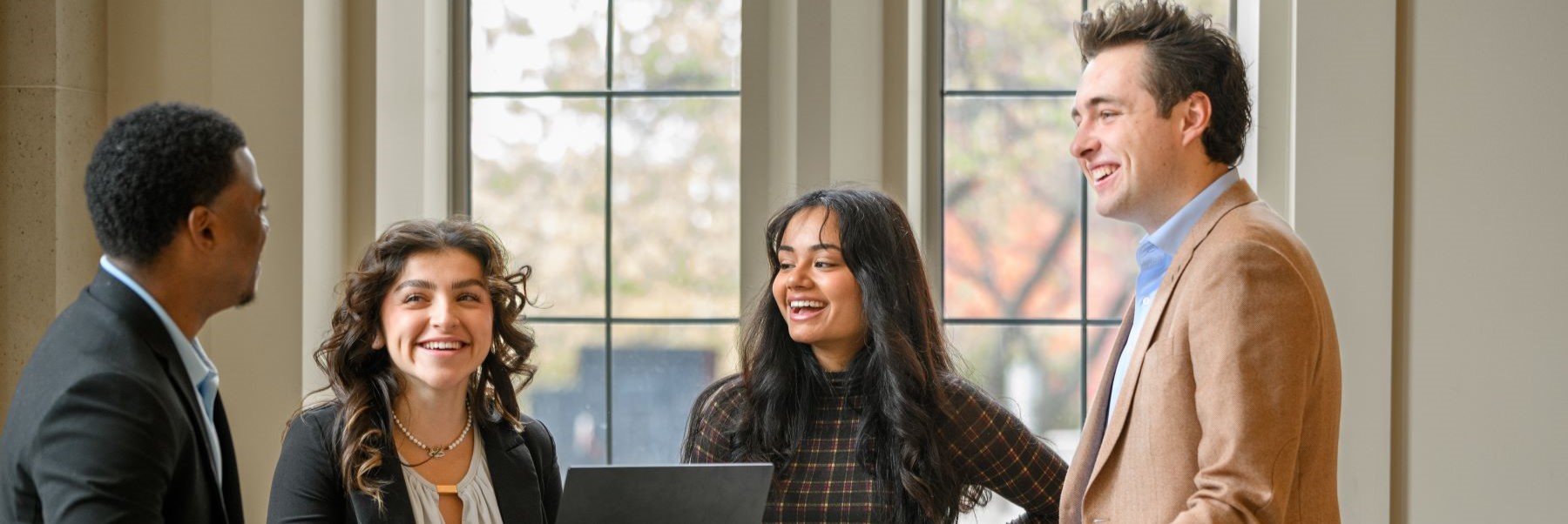 Four people stand inside conversing with two large windows behind them.