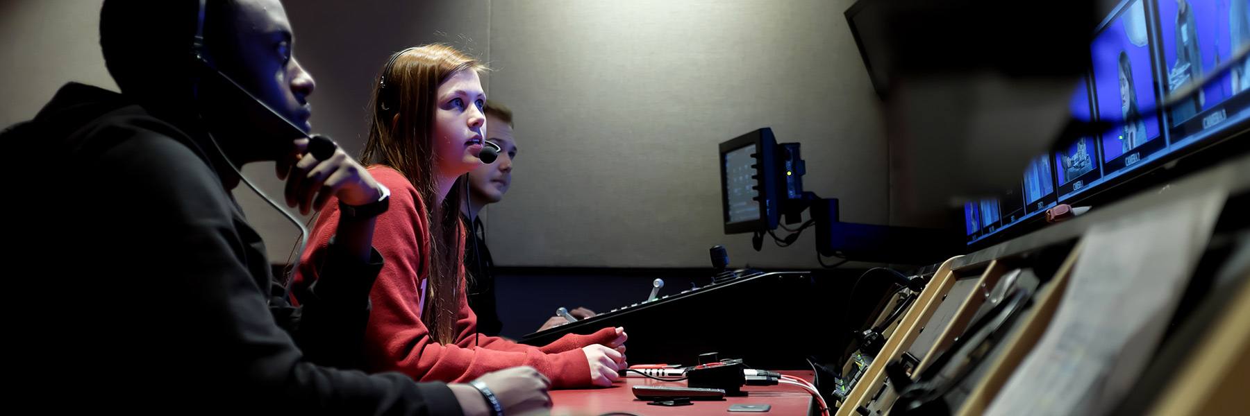 Students sit at a behind-the-scenes TV production station and watch the recorded video on screens.