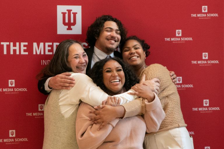 Four people stand in front of a Media School photo backdrop.