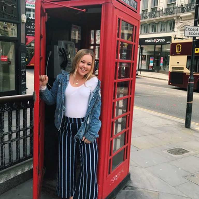 A woman stands in front of a red telephone booth on a city street in London.